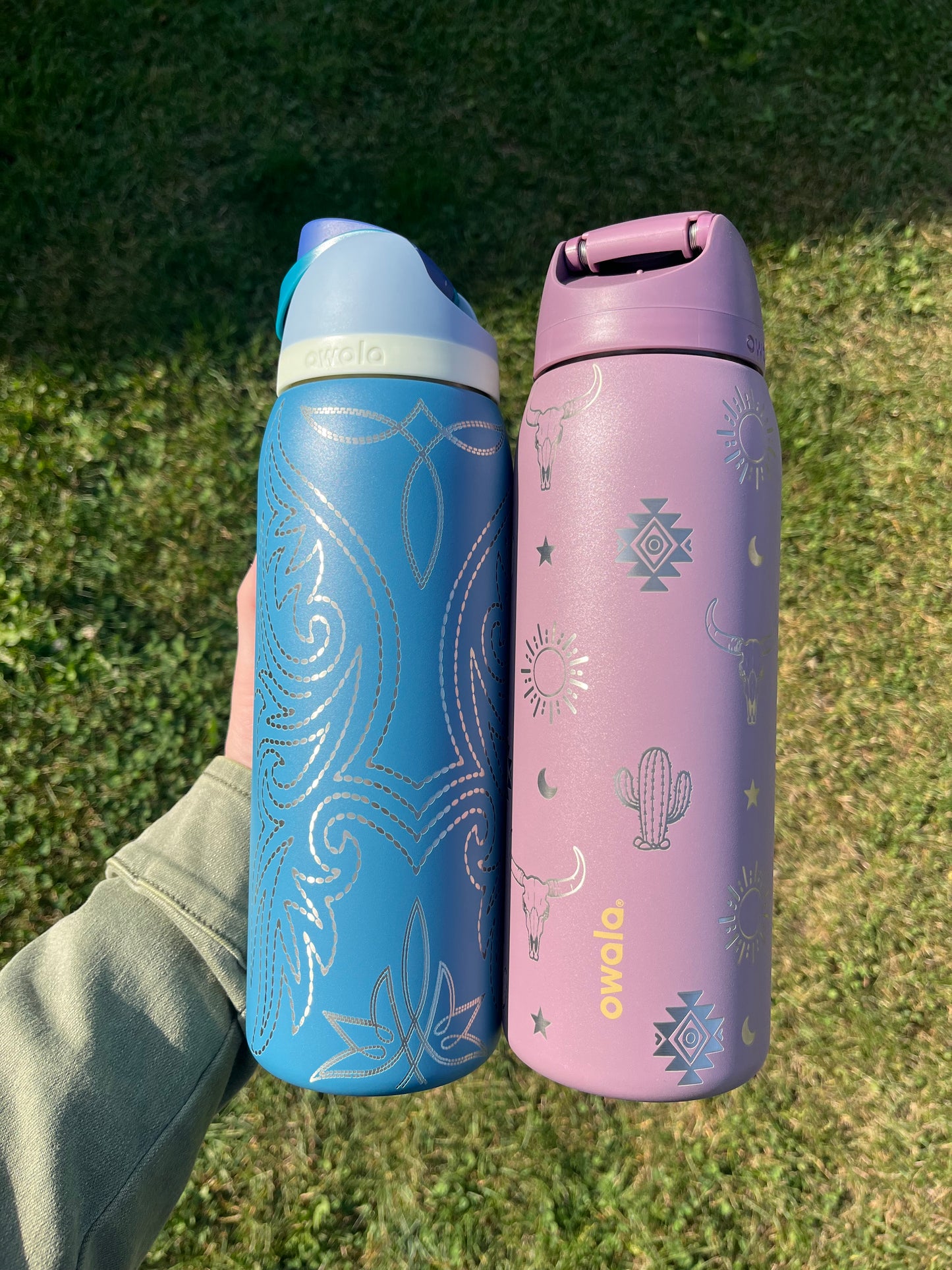 Two insulated bottles, one pink and one blue, held against a grassy background.