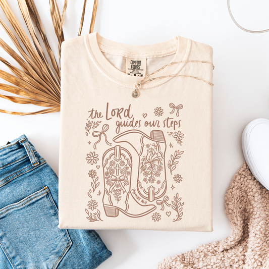 Flat lay of a cream Comfort Colors graphic tee with a western Christian illustration of cowgirl boots filled with florals, crosses, and the phrase ‘The Lord guides our steps