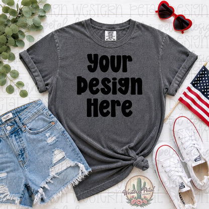Comfort Colors pepper gray t-shirt mockup with distressed denim shorts, white sneakers, patriotic American flag, and heart sunglasses styled in a flat lay for Fourth of July shirt designs.