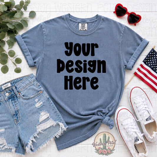 Comfort Colors washed denim blue t-shirt mockup with distressed denim shorts, white sneakers, patriotic American flag, and heart sunglasses styled in a flat lay for Fourth of July shirt designs.
