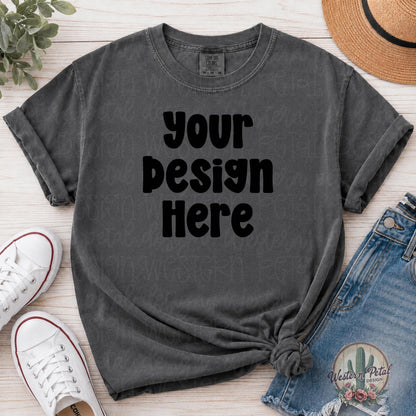 Blank Comfort Colors 1717 pepper color t-shirt mockup with rolled sleeves, front knot styling, denim shorts, white sneakers, straw hat, and eucalyptus on light wood background.