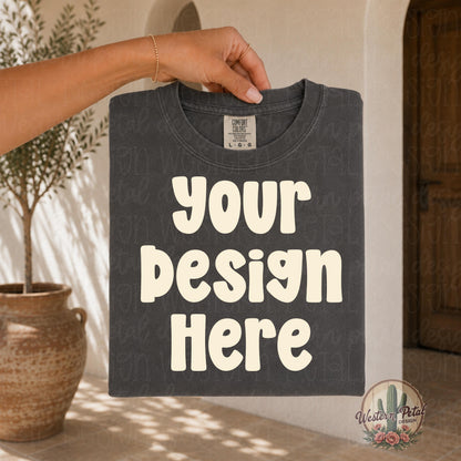 Hand holding a folded Comfort Colors pepper t shirt mockup in a warm neutral boho setting with aesthetic boutique styling and soft natural lighting.