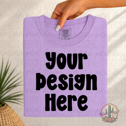 Folded orchid Comfort Colors t-shirt mockup held from the top by a woman’s hand with neutral nails in a soft boho western setting.
