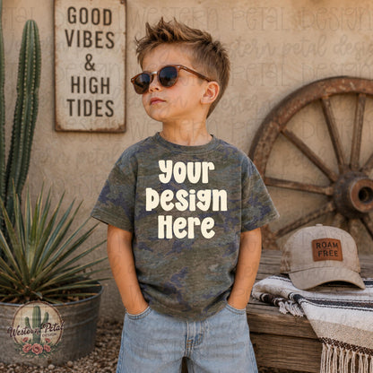 Little boy wearing a vintage camo Rabbit Skins t-shirt mockup with sunglasses in a western desert styled scene with rustic decor and wagon wheel backdrop.