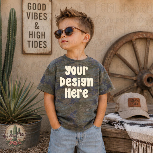 Little boy wearing a vintage camo Rabbit Skins t-shirt mockup with sunglasses in a western desert styled scene with rustic decor and wagon wheel backdrop.