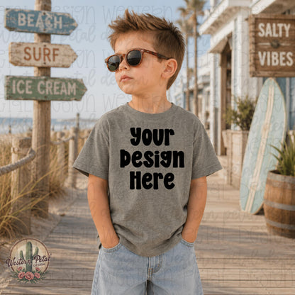 Little boy wearing a Bamboo Blackout Rabbit Skins t-shirt mockup standing on a beach boardwalk with coastal summer decor and surf signs.