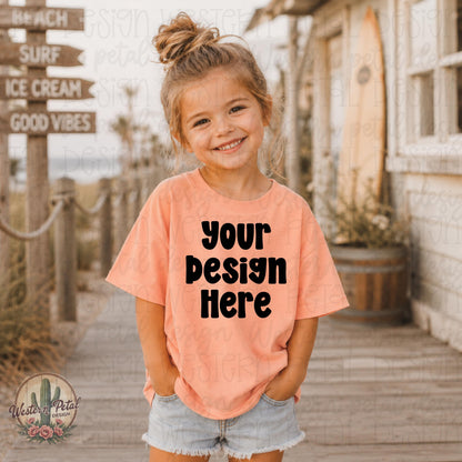 Smiling little girl wearing a peach Rabbit Skins t-shirt mockup on a beach boardwalk with coastal summer decor and surf inspired background.