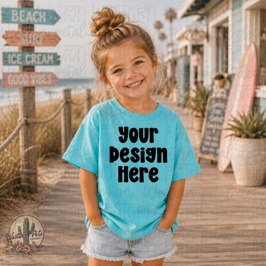 Smiling little girl wearing a turquoise Rabbit Skins t-shirt mockup on a sunny beach boardwalk with colorful coastal summer decor.