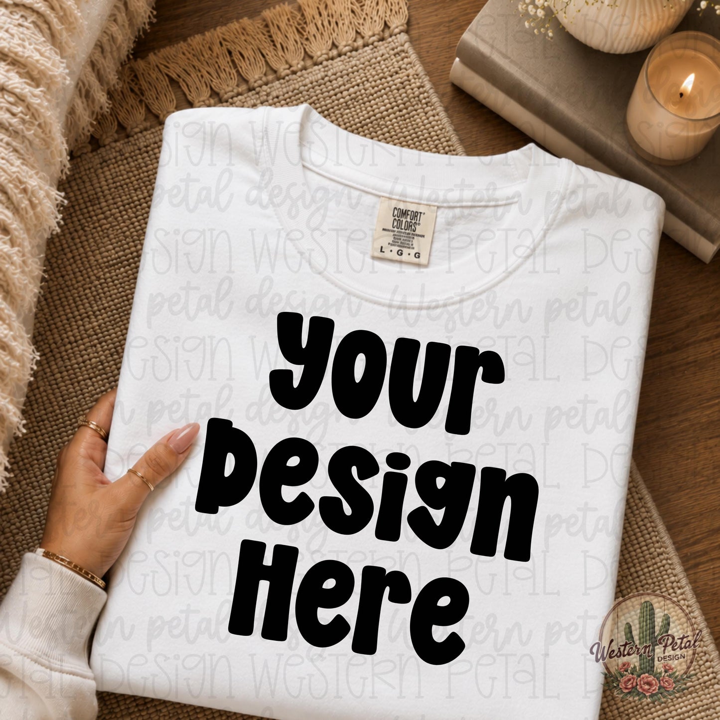 Folded white Comfort Colors t-shirt mockup with a woman’s hand holding the shirt in a cozy neutral boho flat lay setting with candles and textured decor.