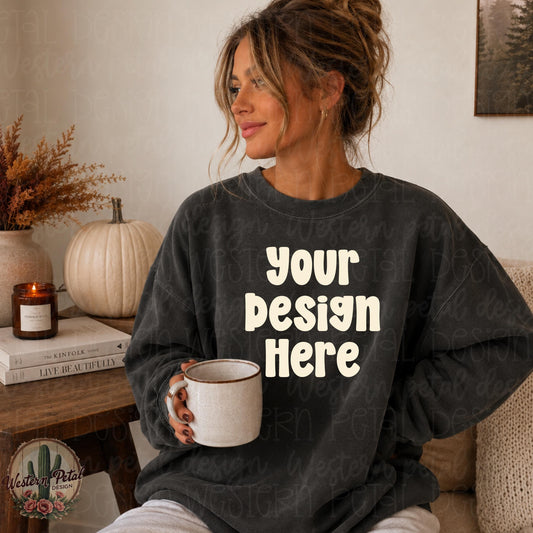 Woman with messy bun wearing a pepper Comfort Colors 1566 crewneck sweatshirt in a cozy neutral fall living room, holding sweatshirt flat for mockup display.