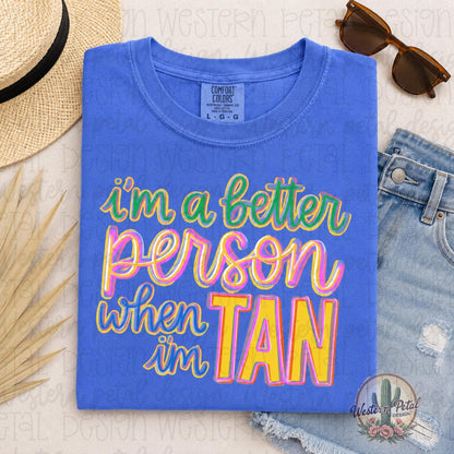 Comfort Colors Flo Blue graphic t-shirt with colorful retro “I’m a Better Person When I’m Tan” design displayed in a styled summer flat lay mockup with denim shorts and sunglasses.