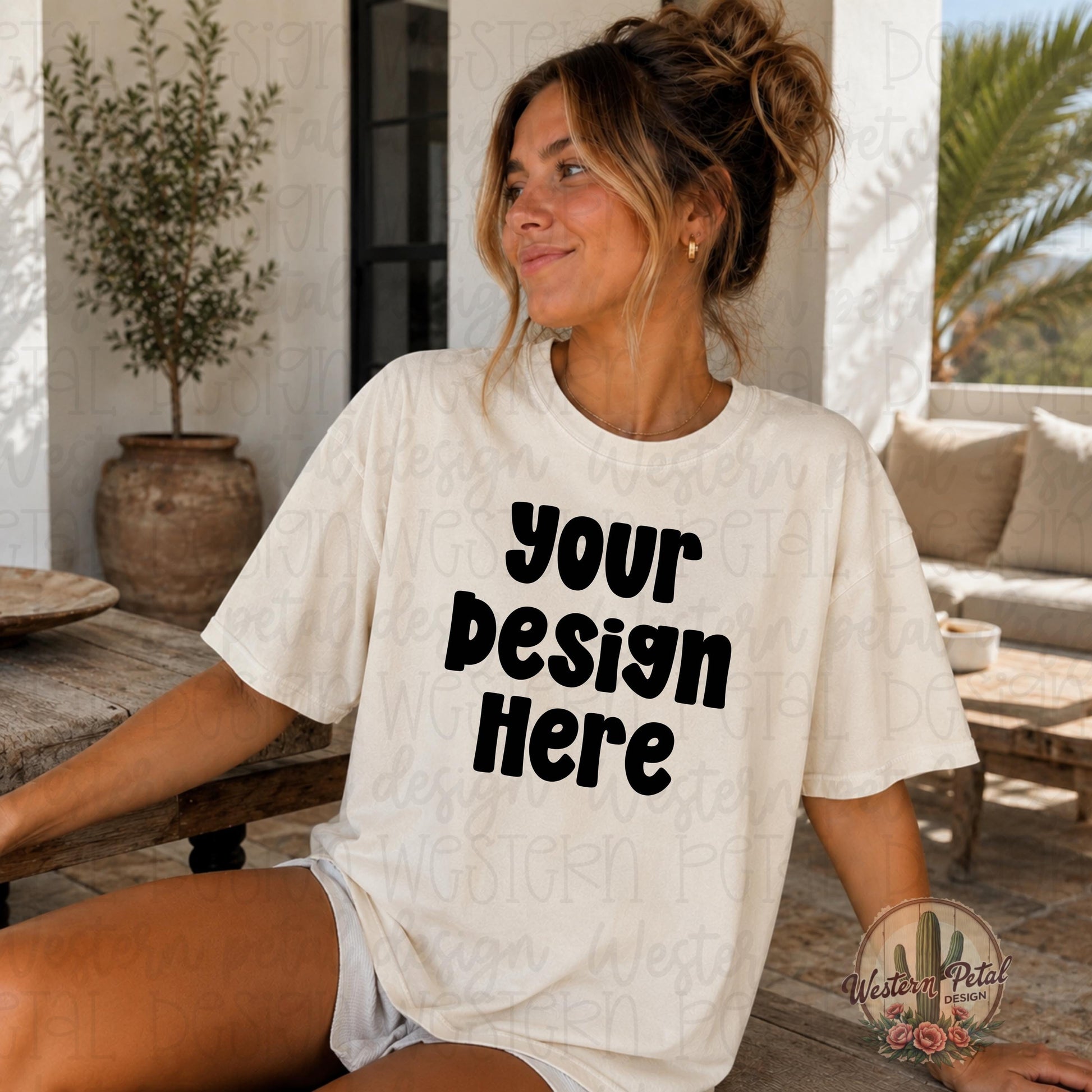 Woman wearing a blank Comfort Colors ivory t shirt sitting outdoors in a warm boho styled patio setting with neutral tones and messy bun hairstyle.