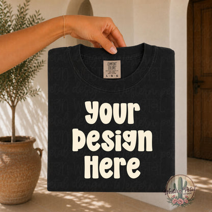 Hand holding a folded Comfort Colors black t shirt mockup in a warm boho styled setting with earthy neutral tones and soft natural lighting.