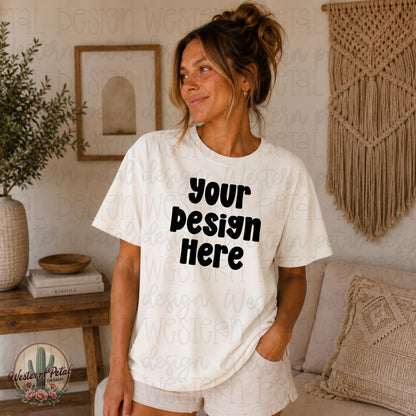 Neutral boho Comfort Colors white mockup featuring a brunette model with a messy bun wearing an oversized white t-shirt in a cozy indoor setting with macrame wall decor and earthy neutral tones.