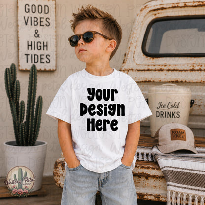 Stylish little boy wearing a white Rabbit Skins t-shirt mockup with sunglasses standing beside a rustic vintage truck and western summer decor.