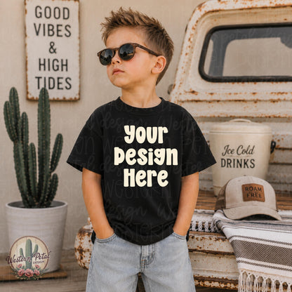 Little boy wearing a black Rabbit Skins t-shirt mockup with sunglasses standing beside a rustic vintage truck and western styled summer decor.