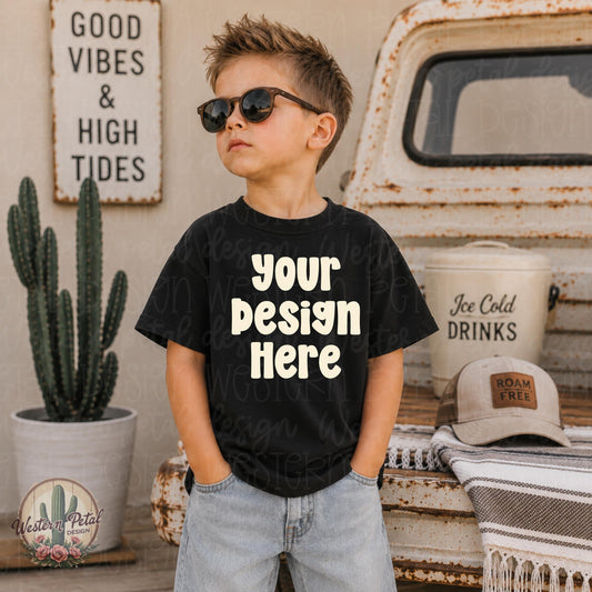 Little boy wearing a black Rabbit Skins t-shirt mockup with sunglasses standing beside a rustic vintage truck and western styled summer decor.