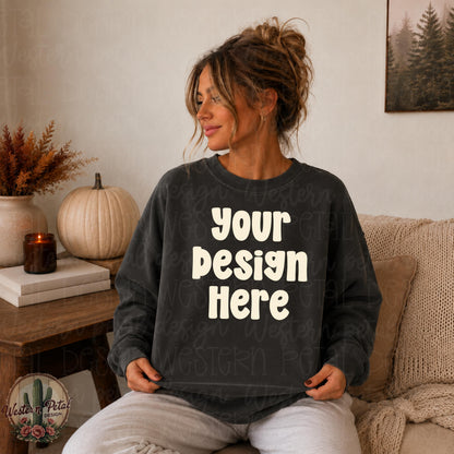 Woman with messy bun wearing a pepper Comfort Colors 1566 crewneck sweatshirt in a cozy neutral fall living room, holding sweatshirt flat for mockup display.