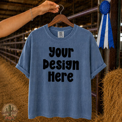 Comfort Colors 1717 Blue Jean t-shirt mockup hanging on wooden hanger in livestock barn with blue ribbon and show ring background.