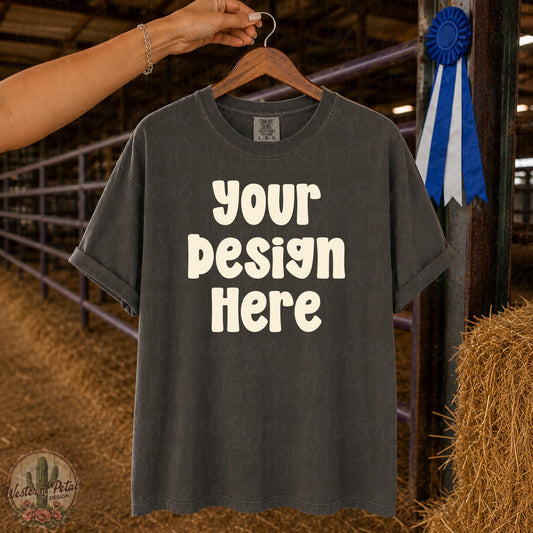 Comfort Colors 1717 Pepper t-shirt mockup hanging on wooden hanger in livestock barn with blue ribbon and show ring background.